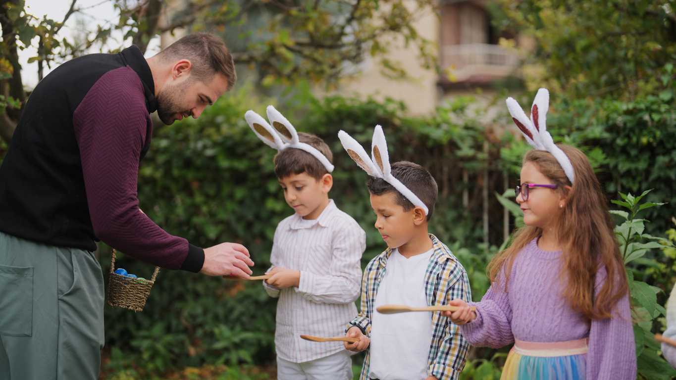 A young man is explaining the rules of egg and spoon race to a group of children outdoors.
