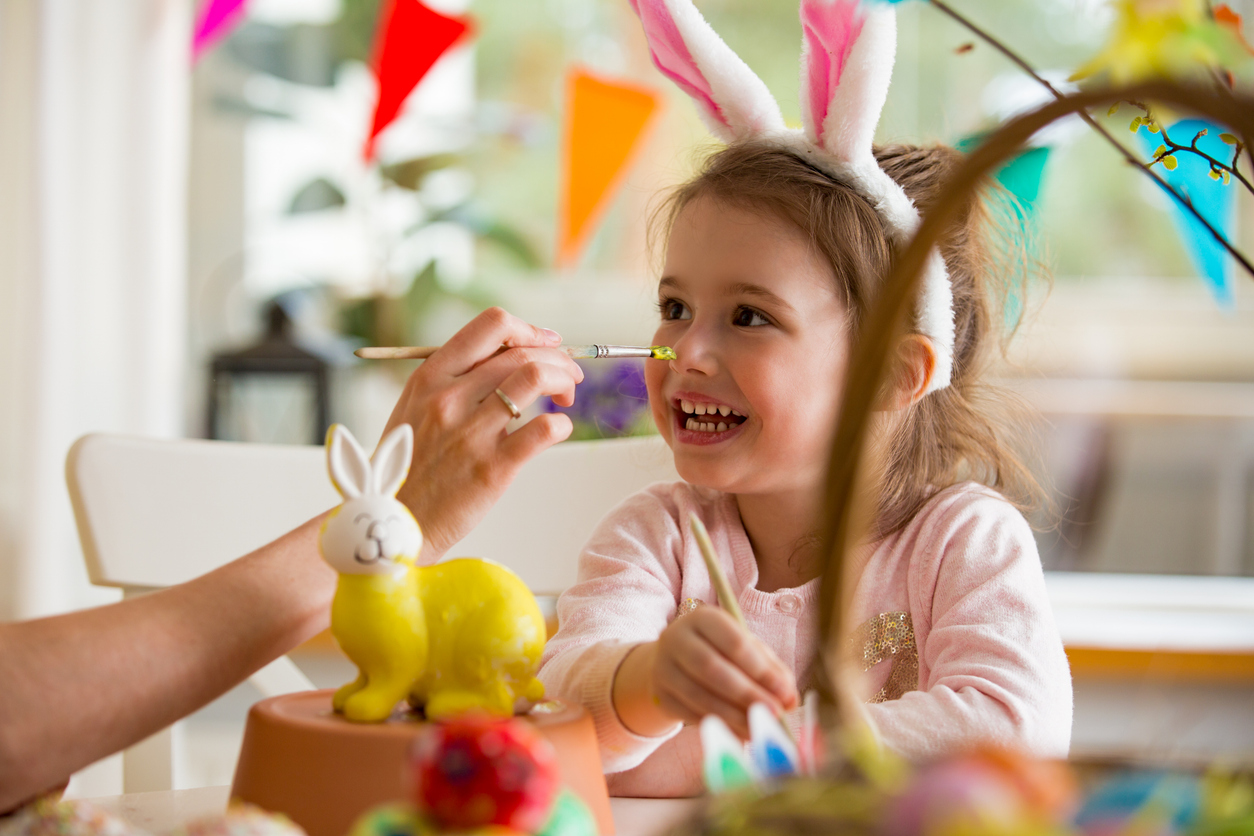 A mother and daughter celebrating Easter, painting bunny with brush in yellow color. Happy family smiling and laughing. Cute little girl in bunny ears preparing the holiday.